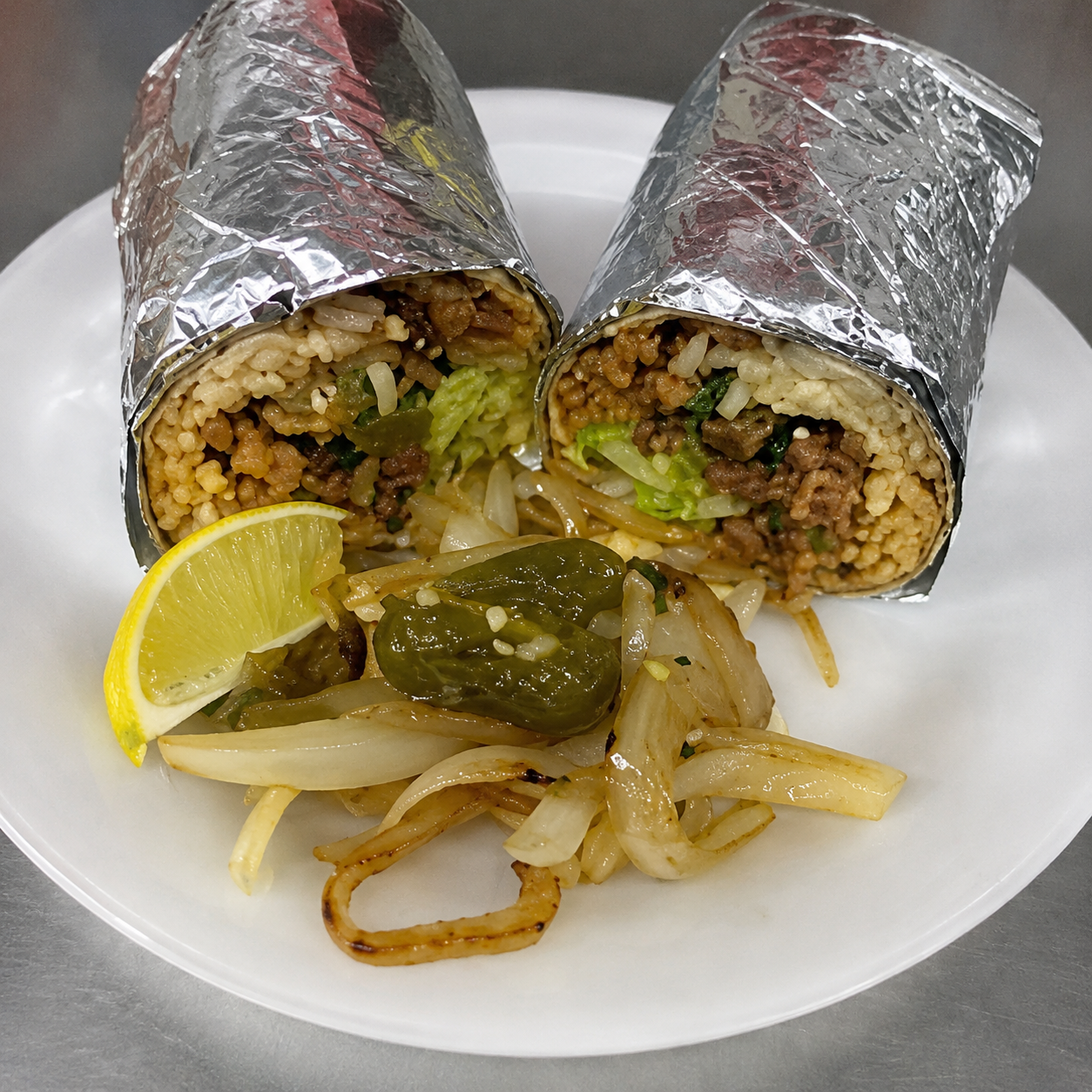 Foil-wrapped burrito cut in half showing rice, meat, avocado, and onion, with chiles toreados and lime on the side